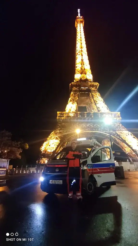 Propozycja: Ratownik medyczny stoi przed ambulansem z otwartymi drzwiami, na tle oświetlonej Wieży Eiffla w Paryżu. Międzynarodowy transport medyczny Włocławek.